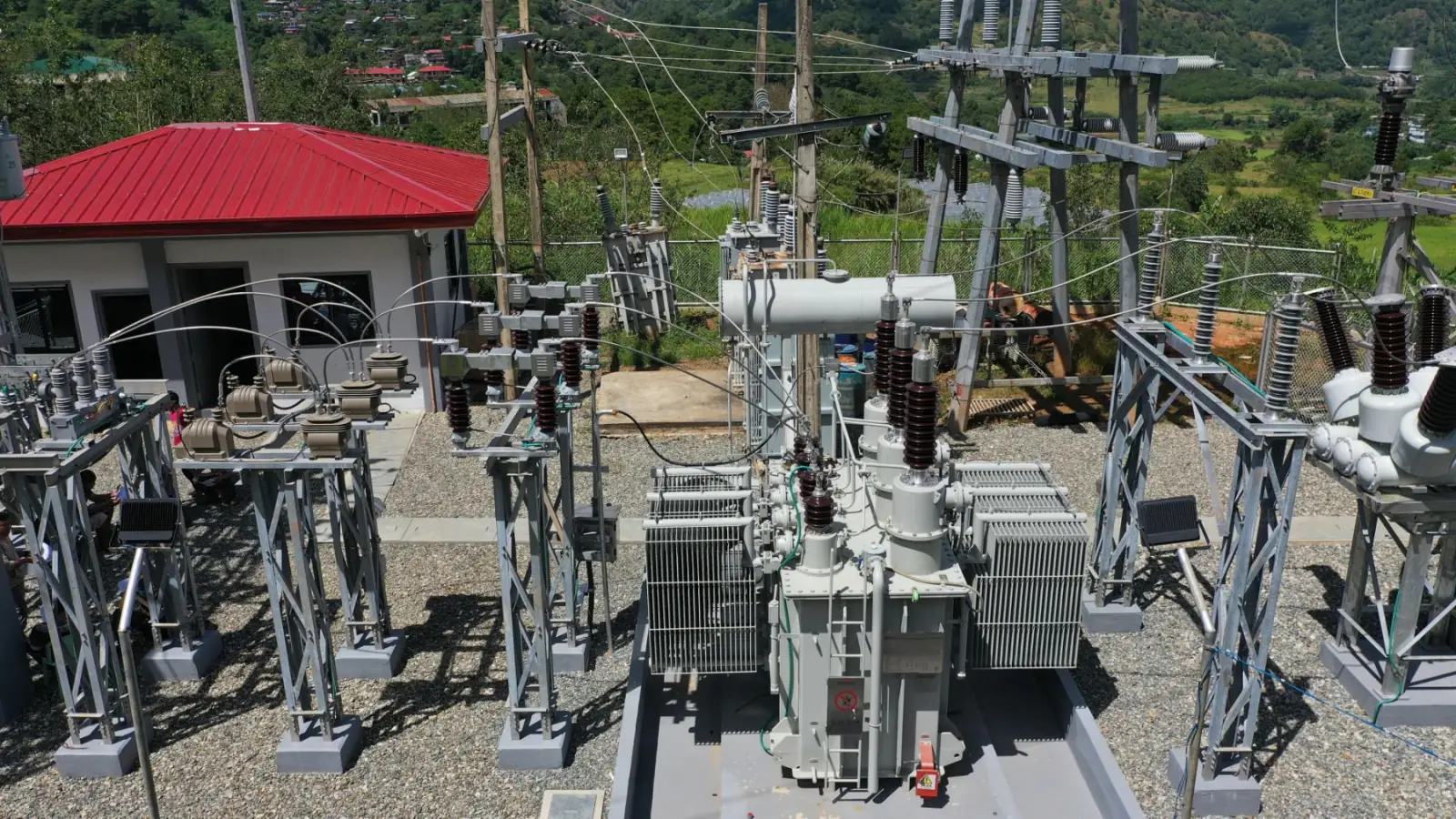 Substation with a power transformer and red-roofed control house against a green mountain backdrop